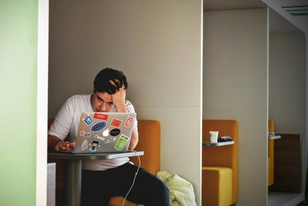 man wearing white top using MacBook. He looks frustrated, supporting his head with his hand, his fingers in his hair.